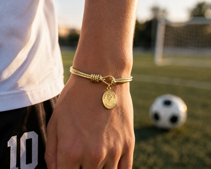 A gold-toned cord bracelet with a knotted detail and a round coin charm featuring a soccer ball design, worn on a person's wrist in a sunny outdoor soccer field setting. Carlo Acutis 