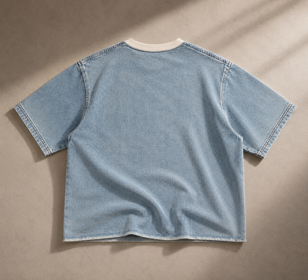 A light blue denim short-sleeve t-shirt shown from the back, laid flat on a textured beige surface with soft diagonal shadows.