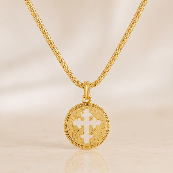 A gold chain necklace with a round gold pendant featuring a white cross in the center, displayed on a light beige background.