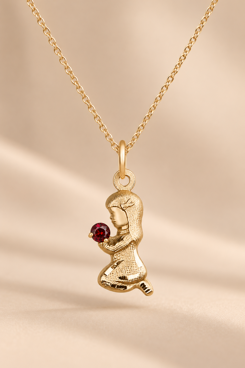 A gold pendant necklace featuring a kneeling figure holding a small round red gemstone, displayed on a soft beige background.