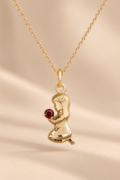 A gold pendant necklace featuring a kneeling figure holding a small round red gemstone, displayed on a soft beige background.