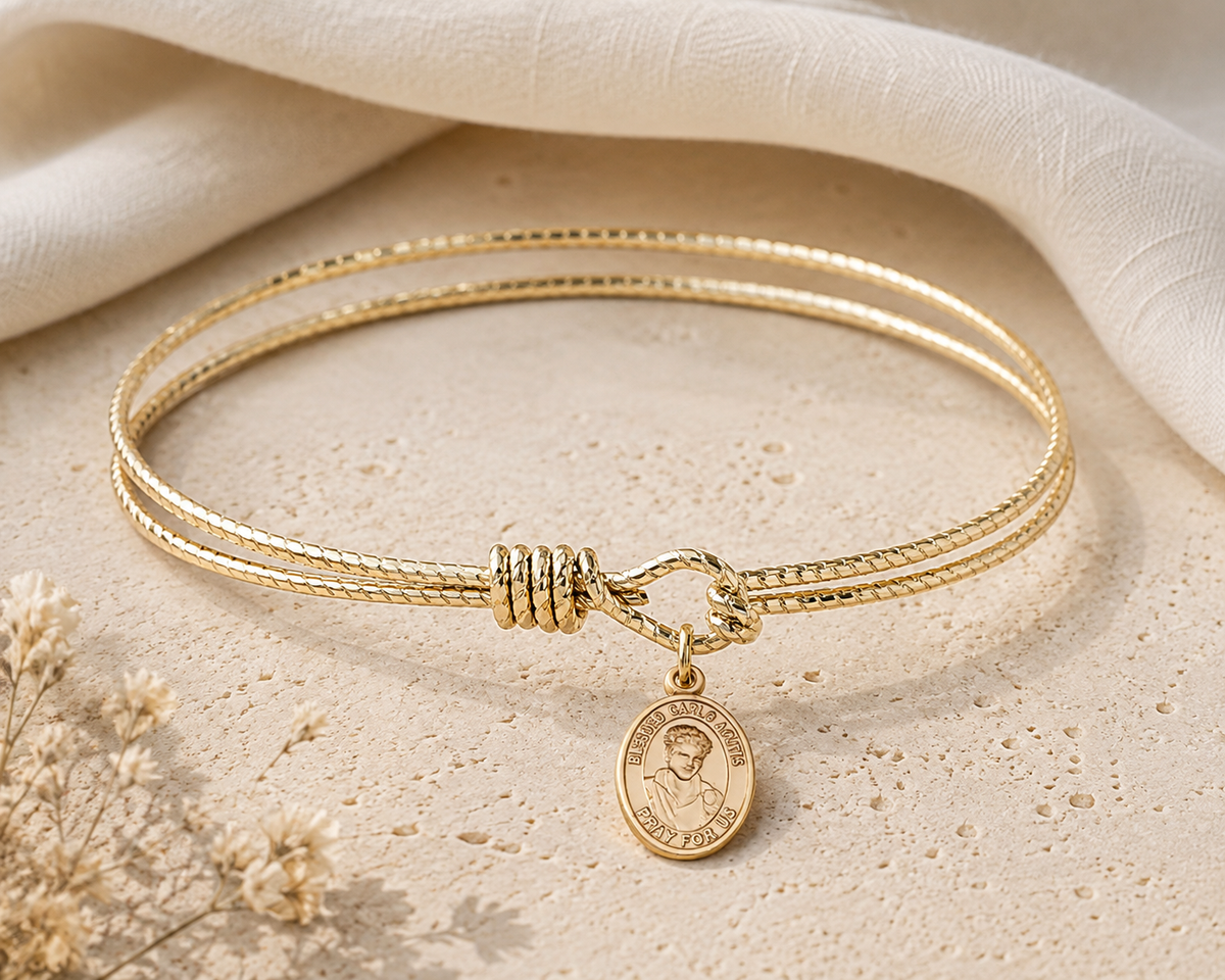 A gold double-strand rope bangle bracelet with a small round Saint Patrick medallion charm, displayed on a textured beige stone surface with a draped cream fabric background.