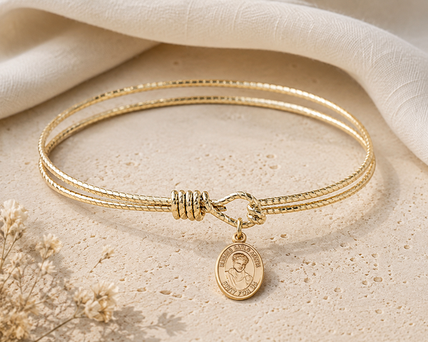 A gold double-strand rope bangle bracelet with a small round Saint Patrick medallion charm, displayed on a textured beige stone surface with a draped cream fabric background.