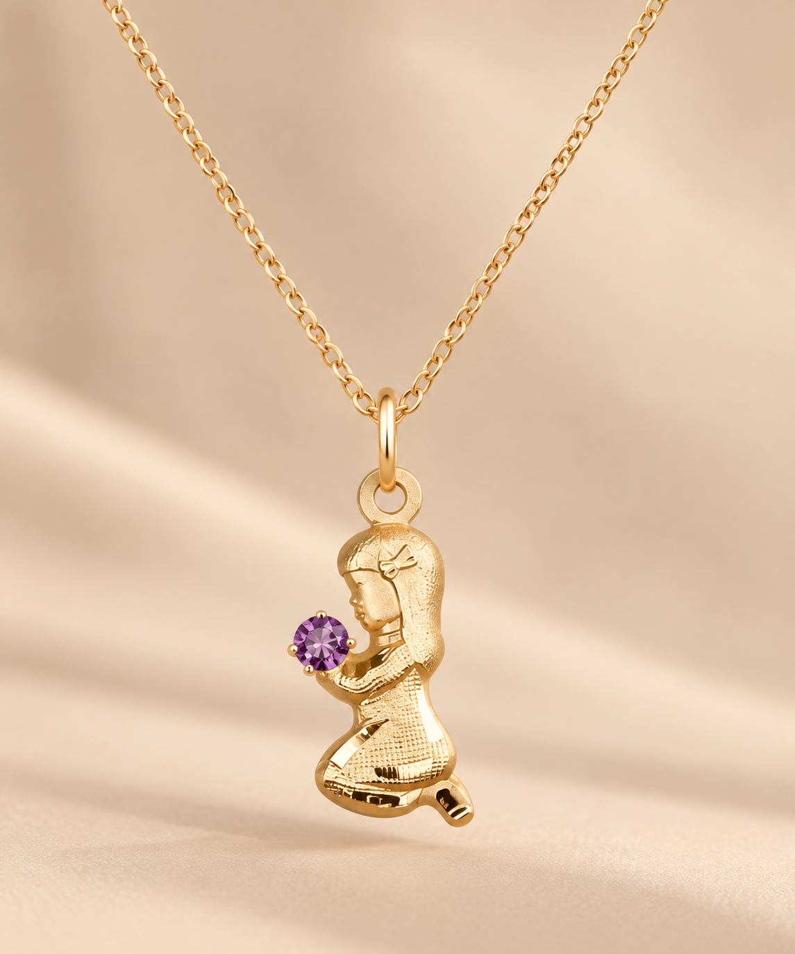 A gold pendant necklace featuring a kneeling girl holding a small purple gemstone, displayed on a soft beige satin background.