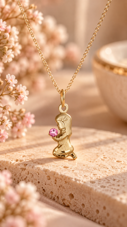 A gold-tone kneeling figure pendant necklace holding a small pink gemstone, displayed on a textured stone surface with soft pink flowers and a warm neutral background.