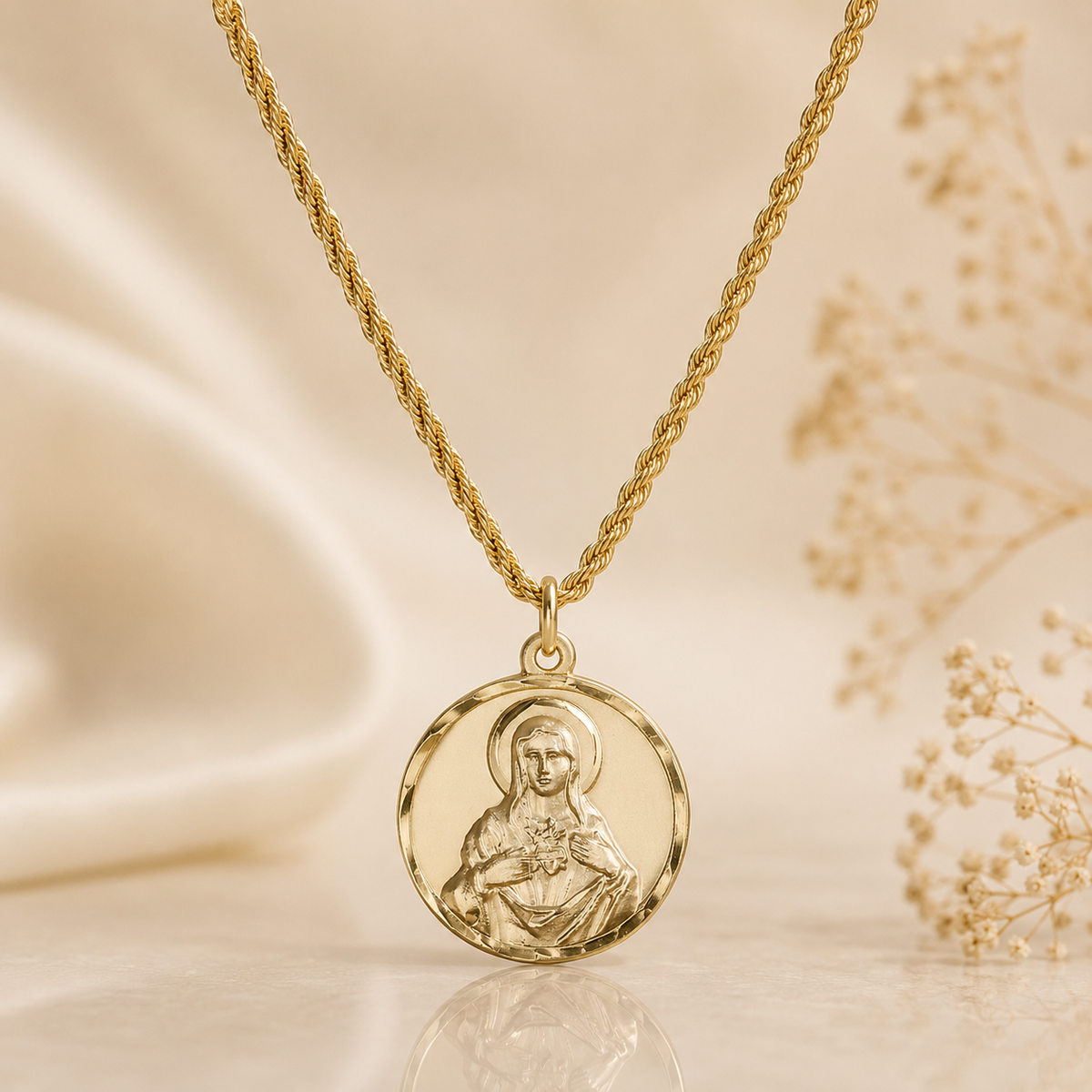 A gold necklace with a round medallion pendant featuring an embossed religious figure, displayed on a soft beige satin surface with dried flowers in the background.