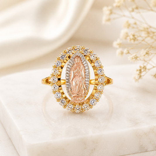 A gold and rose gold Miraculous Medal ring with clear gemstones, displayed on a white marble block with soft cream fabric and baby's breath flowers in the background.