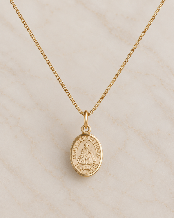 A gold chain necklace with an oval religious pendant featuring a saint's depiction and engraved text, displayed on a light beige marble surface.