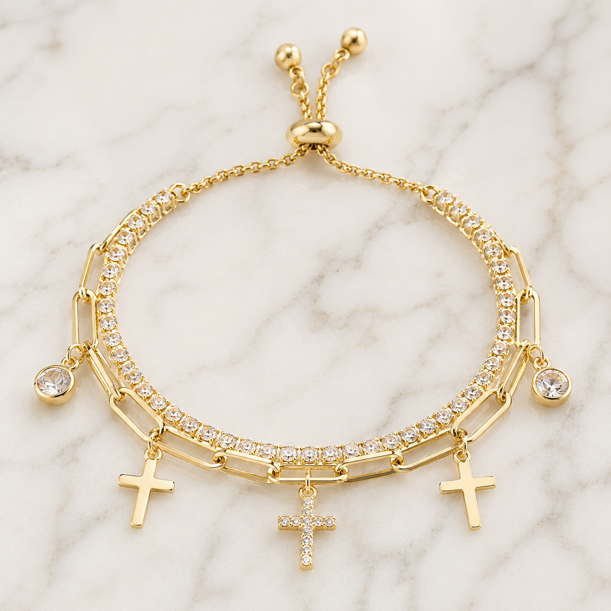 A gold-tone multi-strand bracelet featuring three rows of clear crystals, three dangling cross charms, and a sliding clasp, displayed on a white marble background.