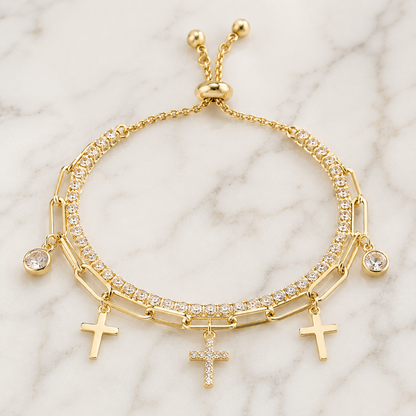 A gold-tone multi-strand bracelet featuring three rows of clear crystals, three dangling cross charms, and a sliding clasp, displayed on a white marble background.