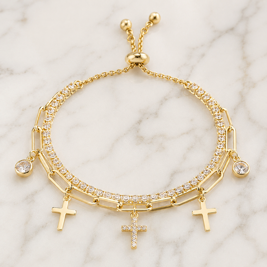 A gold-tone multi-strand bracelet featuring three rows of clear crystals, three dangling cross charms, and a sliding clasp, displayed on a white marble background.