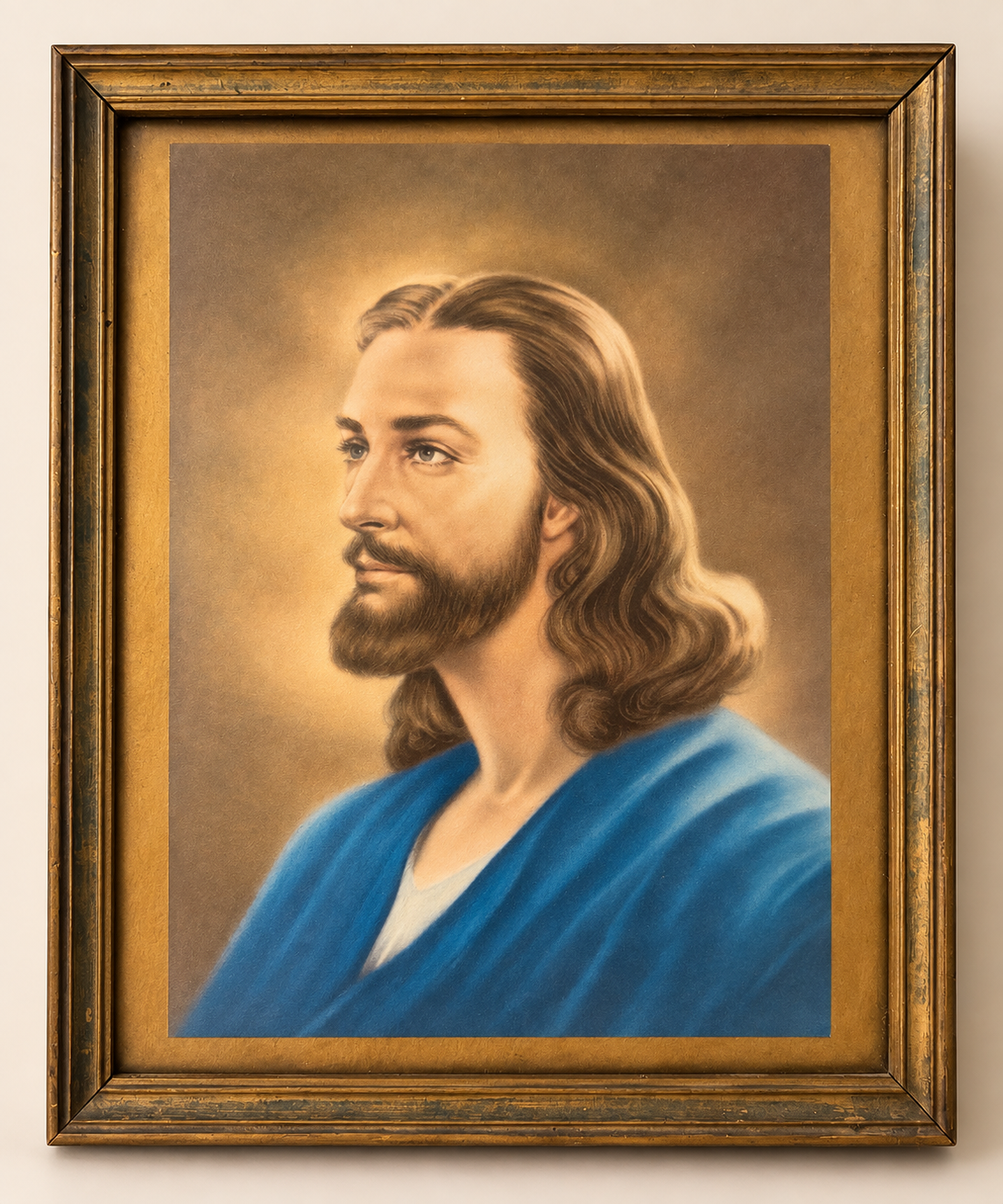 A framed religious portrait painting of Jesus Christ with long brown hair, a beard, and a blue robe, set in a distressed gold frame against a light beige wall.